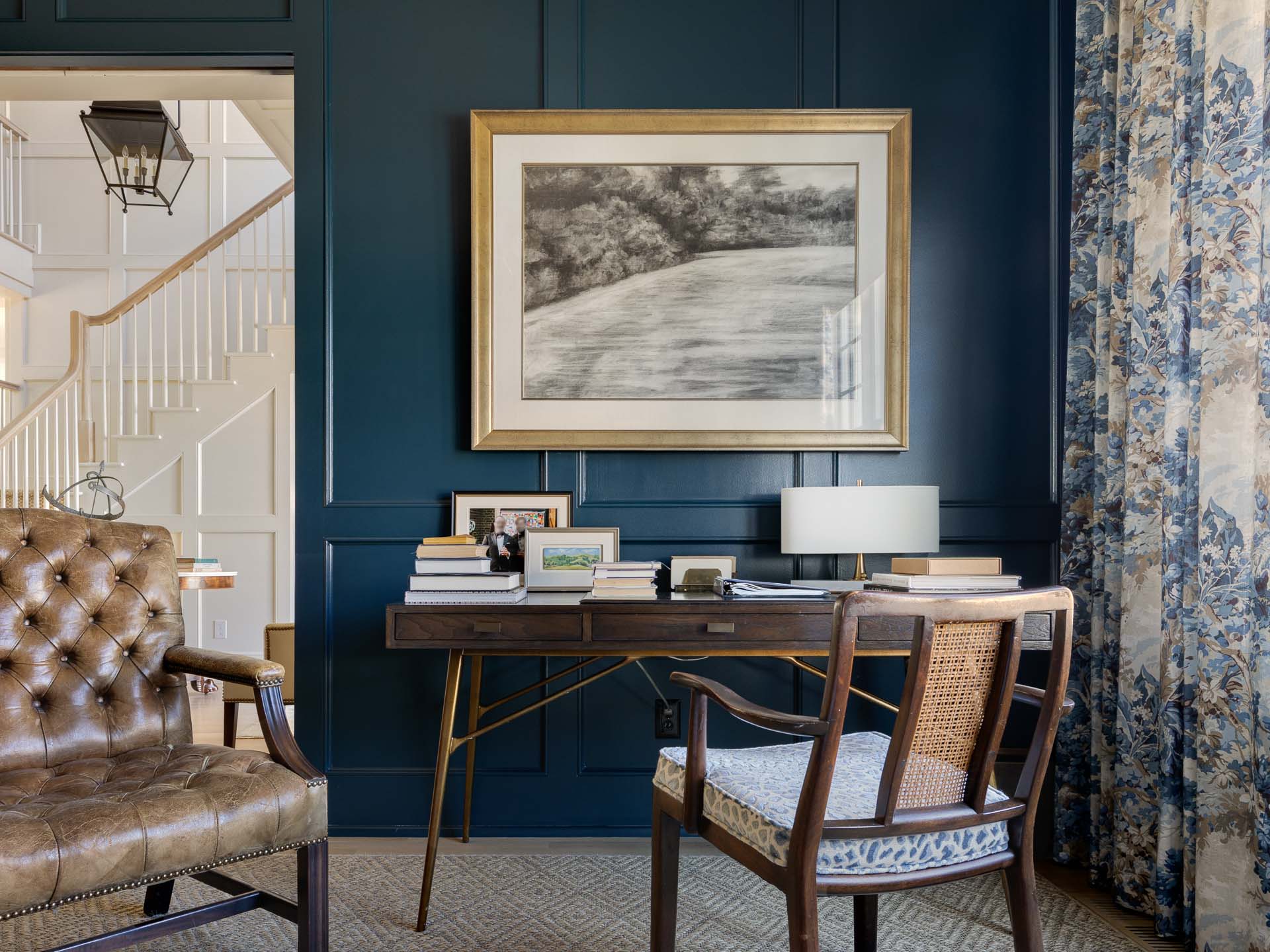 A cozy dark blue office with a mid-century modern wood desk and floral curtains. A large black and white drawing framed in gold sits over the desk the image was captured by the best photographer in Raleigh, North Carolina.